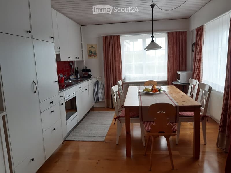 Kitchen with dining area, wooden table and chairs, white cabinets, wooden floor, window with curtains, microwave on the countertop