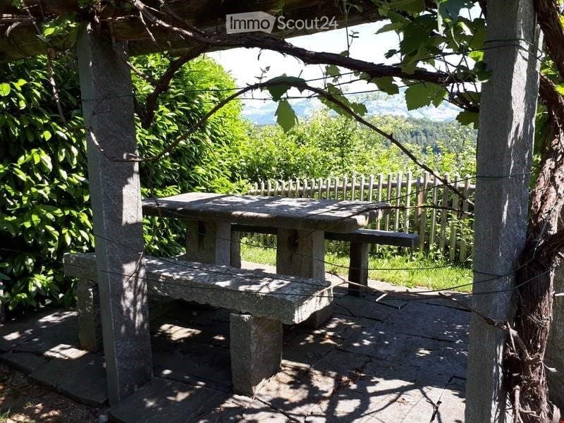 Stone benches under a pergola, stone table, wire fencing, surrounded by greenery, mountain in the distance.
