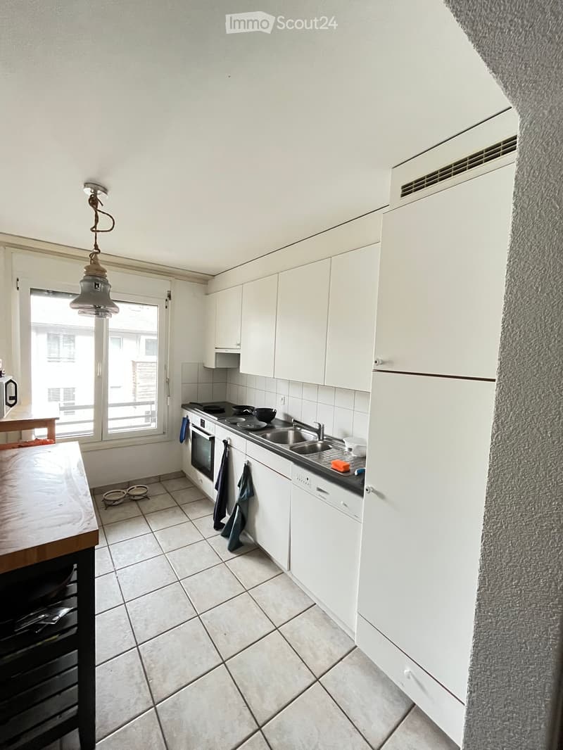 Modern kitchen with white cabinets, tiled floor, and stainless steel appliances including an oven, sink, and dishwasher. A small table is visible near the window.