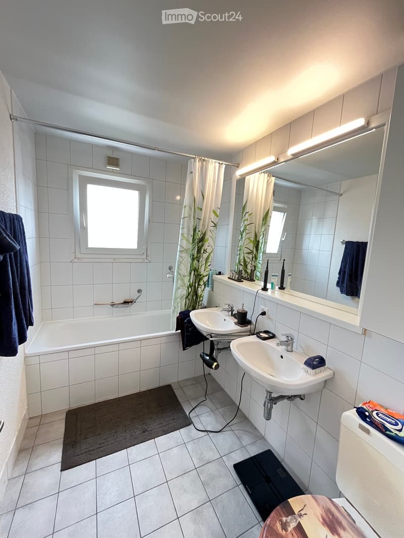 Modern bathroom with white tiles, bathtub, double sink, large mirror, and shower area with a curtain. Includes towels, a hair dryer, and personal care items on the sink counter.