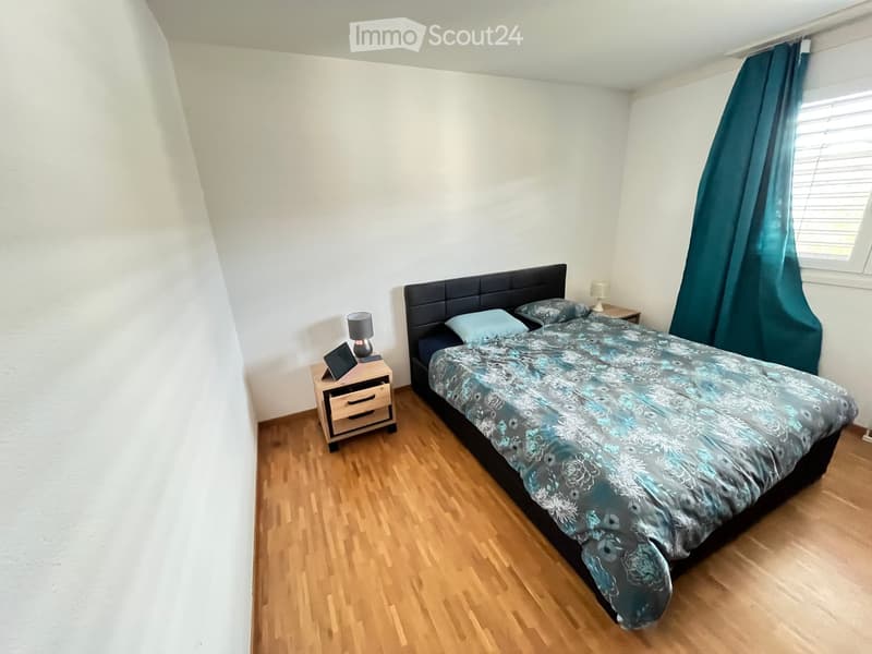 Cozy bedroom with wooden flooring, a large bed with a patterned duvet, two nightstands with lamps, and a small table with a screen on it.