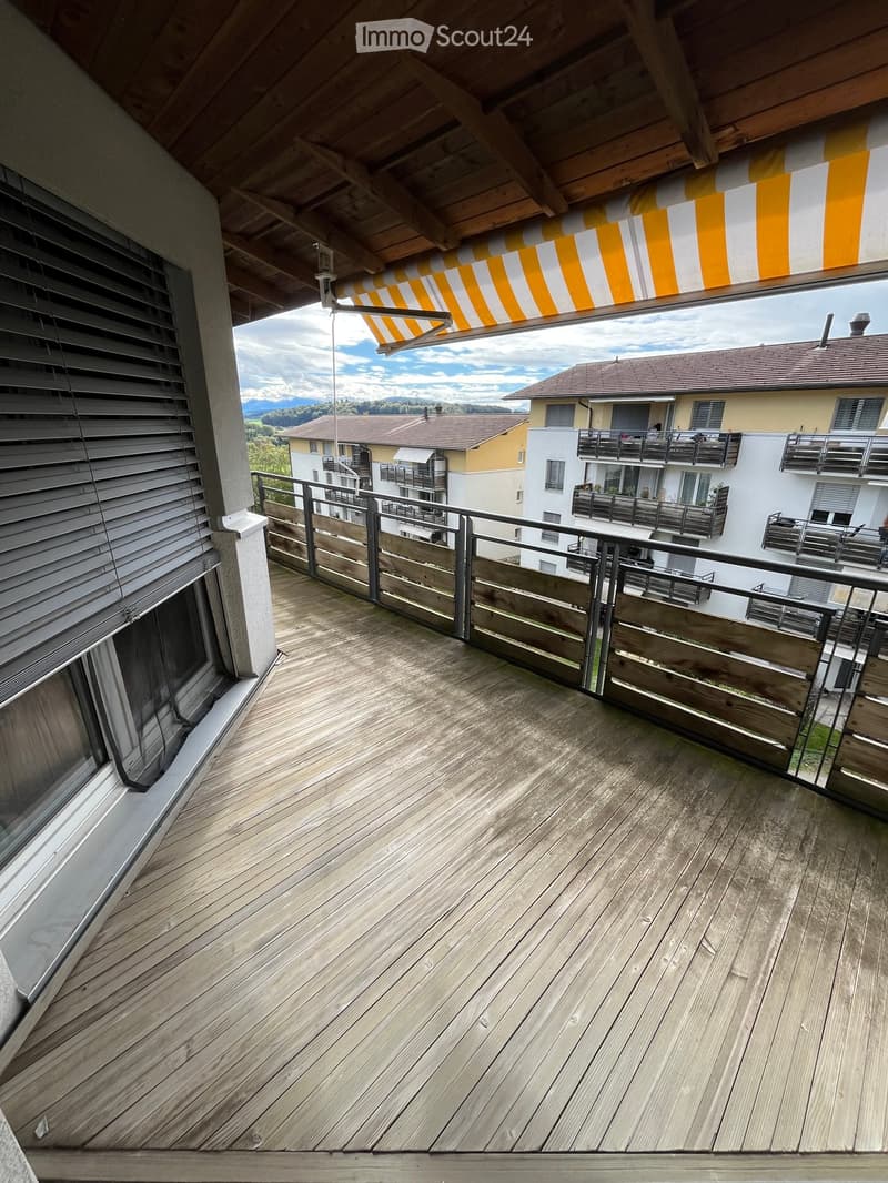 Wooden balcony with metal railing, overlooking a residential area with multiple buildings and a scenic mountain backdrop.