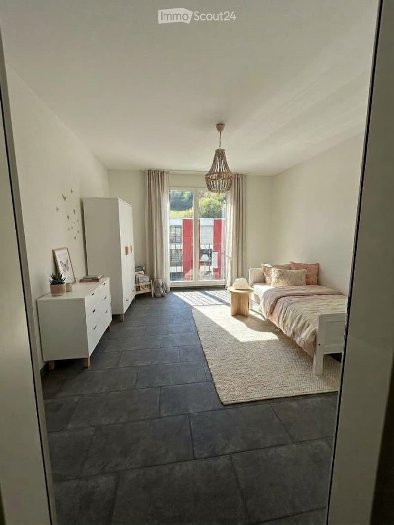 Bright bedroom with grey tiled floor, white walls, large window with view, bed with pillows and blanket, nightstand, chair, wardrobe, small table with potted plant, picture frame on dresser.