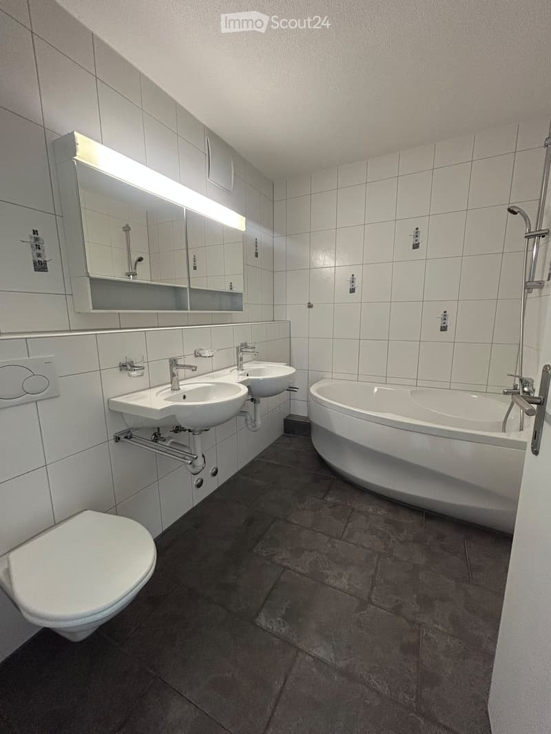 Modern bathroom with white tiled walls, a toilet, two sinks, a bathtub, and a large mirror with built-in lighting.