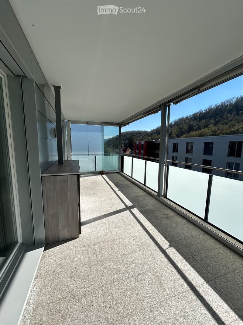 Modern balcony with large windows, tiled floor, and a small wooden table near the wall. Offers a scenic view of the surrounding area.