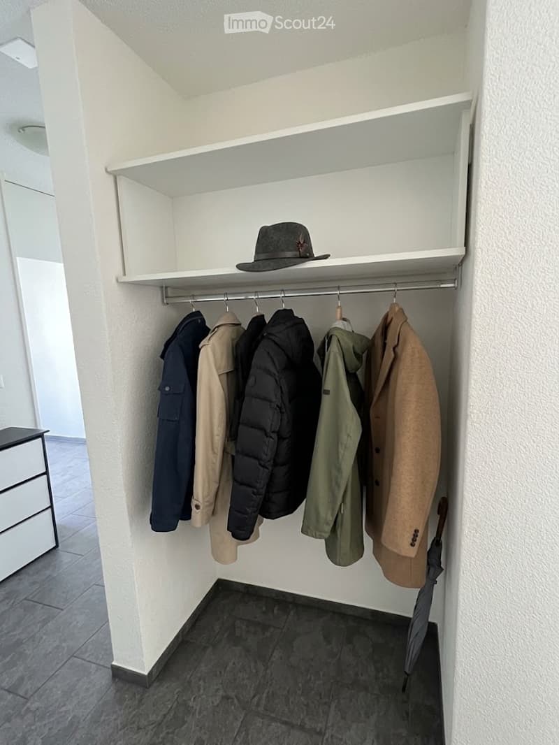 A small, well-lit closet with white walls and a tiled floor. It features a built-in shelf holding a hat and a rod with several coats hanging. An umbrella is placed on the floor near the rod.