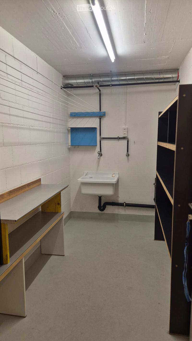 Empty room with white brick walls, a concrete floor, a sink, and exposed pipes. There is a wooden shelf on the left side and a black cabinet on the right side.