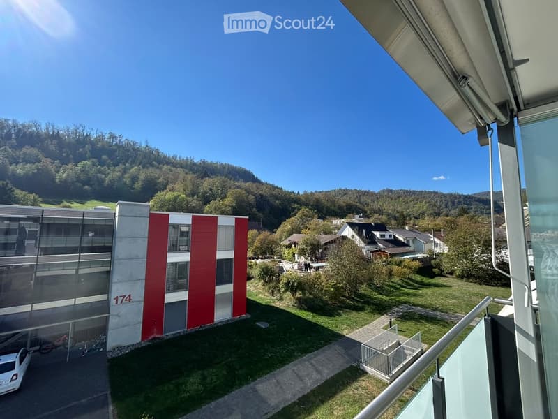 Modern building with red and white facade, number 174 visible, surrounded by green lawn and trees, with a view of a hilly landscape in the background.