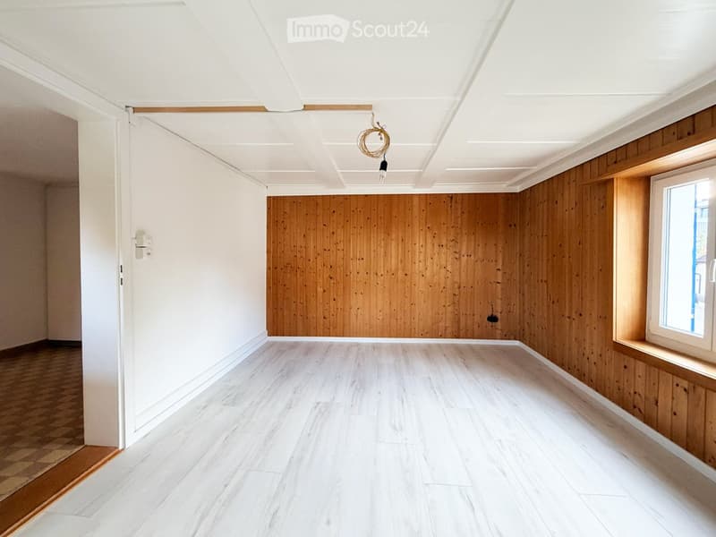 Empty room with white walls, wooden paneling on one wall, white ceiling, and light-colored wooden flooring.
