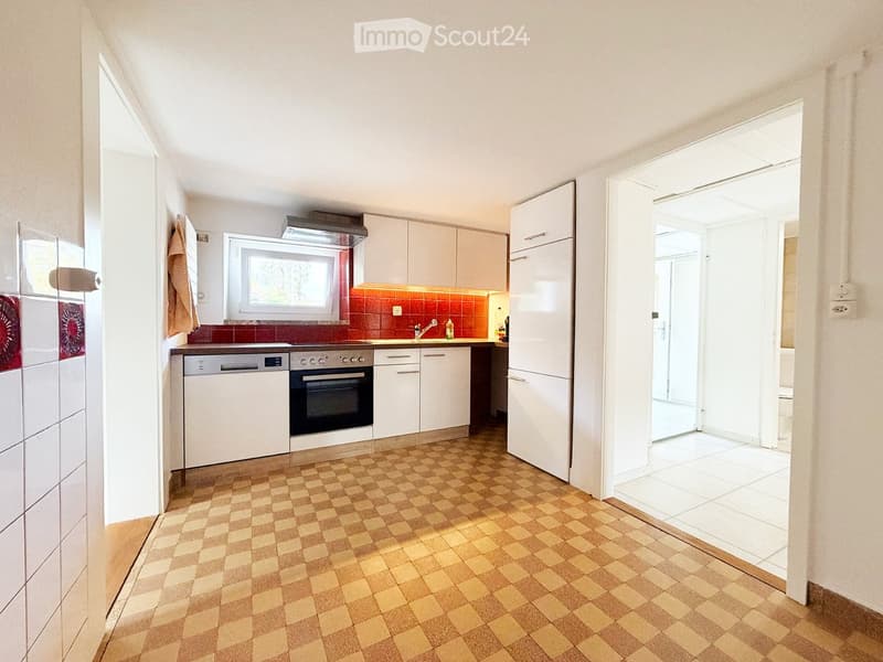 Modern kitchen with white cabinets, a dishwasher, oven, refrigerator, and a window above the sink. The floor is tiled in a checkered pattern, and the backsplash is red.