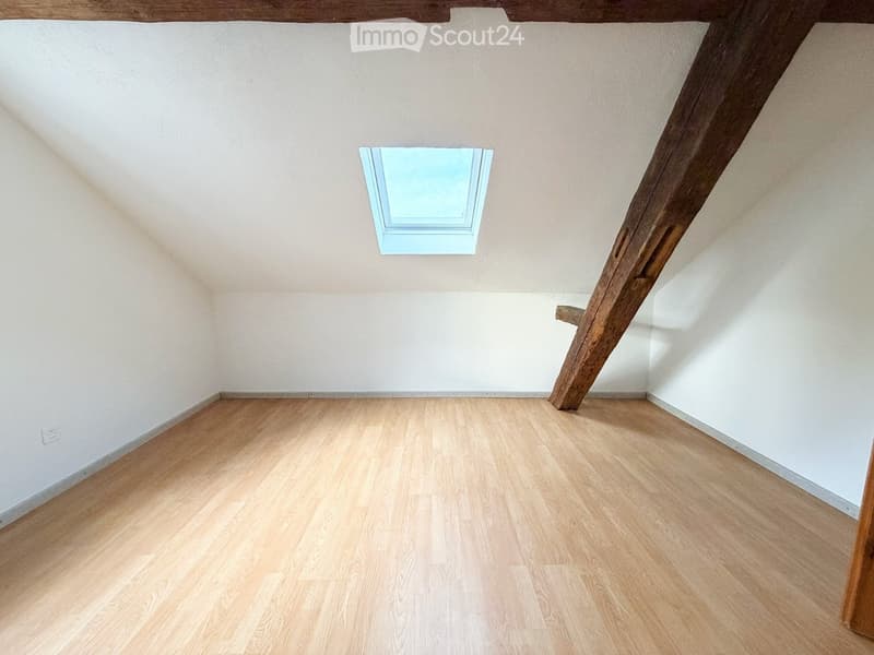 Empty attic room with wooden floor, white walls, and exposed wooden beams. A skylight window provides natural light.