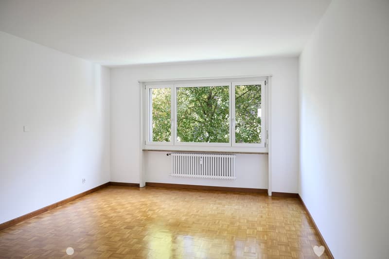 Empty room with white walls, wooden floor, large window with white frame, radiator below window, and visible electrical outlet on wall.