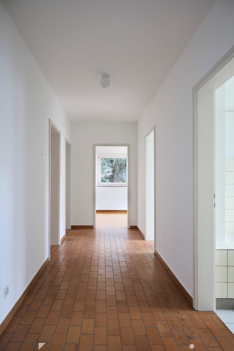 A long, narrow hallway with white walls, wooden tiled flooring, and several doors leading to other rooms. The ceiling is white with a single light fixture.