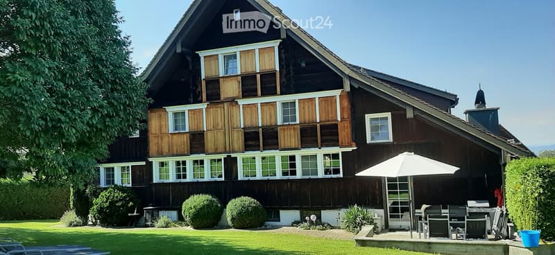 Traditional two-story wooden house with white-framed windows and shutters, surrounded by a well-maintained lawn and garden, with outdoor seating area and umbrella.