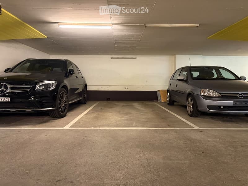 Underground parking area with two parked cars, concrete floor, white walls, and fluorescent lighting.