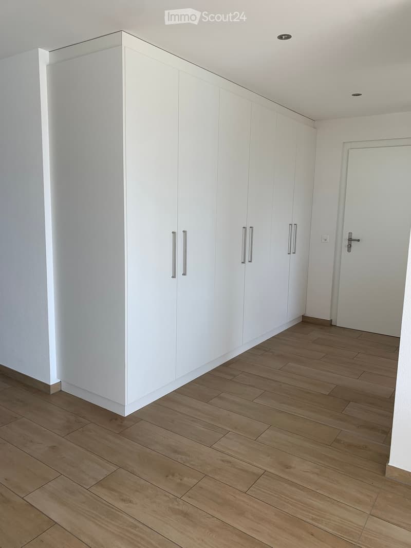 Empty room with light wooden flooring and white built-in wardrobes along one wall. The space is clean and unfurnished, with a white door visible on the right side.