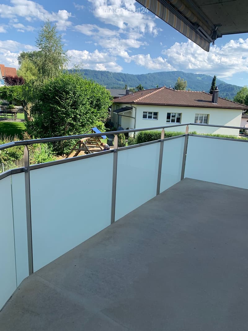 Balcony with white walls and metal railing, overlooking a garden and a house with a tiled roof and multiple windows.