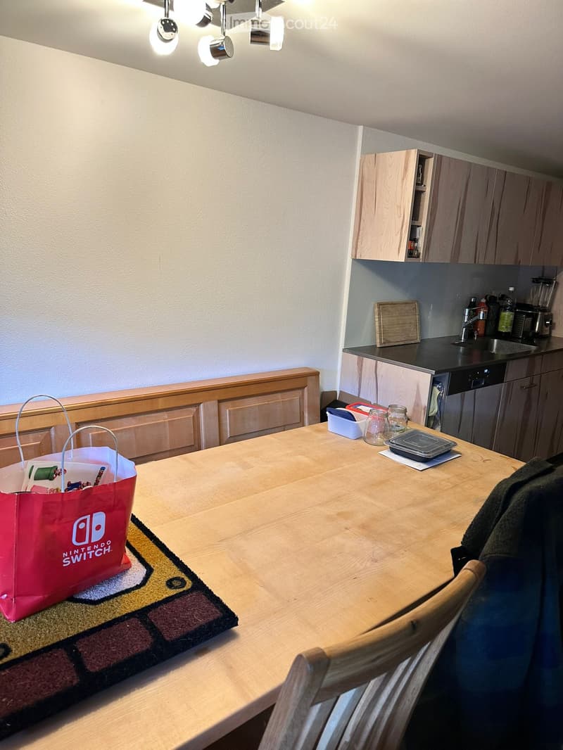 Modern kitchen with wooden table, chairs, and various appliances including a dishwasher, sink, and blender. A Nintendo Switch bag is placed on the table.