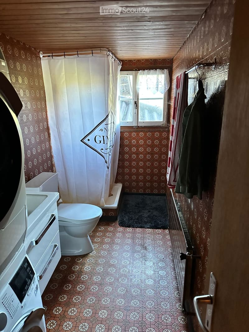 Cozy bathroom with patterned tiles, a toilet, sink, bathtub, and a coat rack with jackets. The space features a small window with a view and a shower curtain.