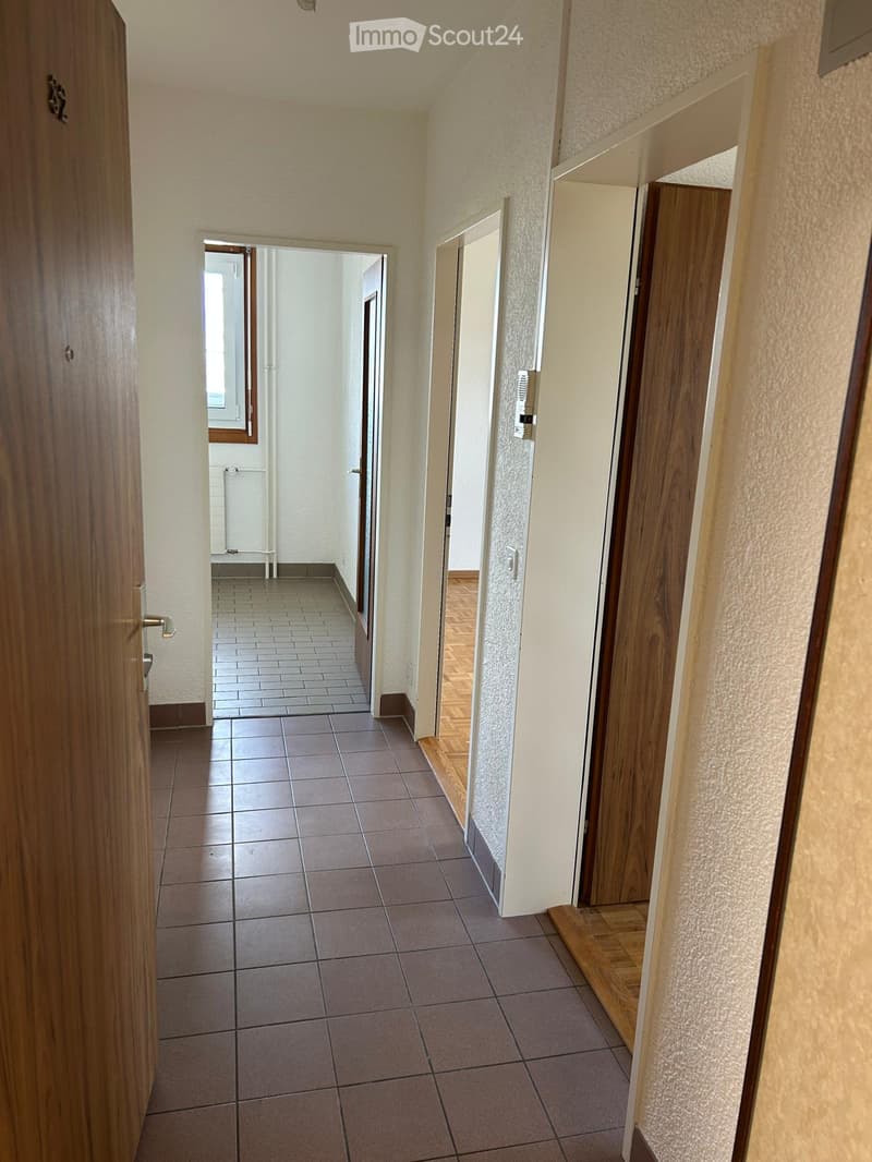 A long hallway with tiled flooring, wooden doors, and white walls. The hallway leads to another room with a window and a radiator visible in the background.