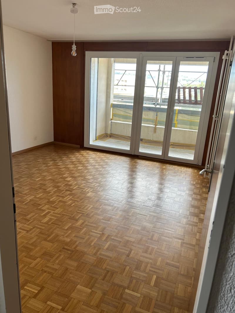 Empty room with wooden herringbone flooring, large sliding glass doors leading to a balcony, and a visible light fixture on the ceiling.