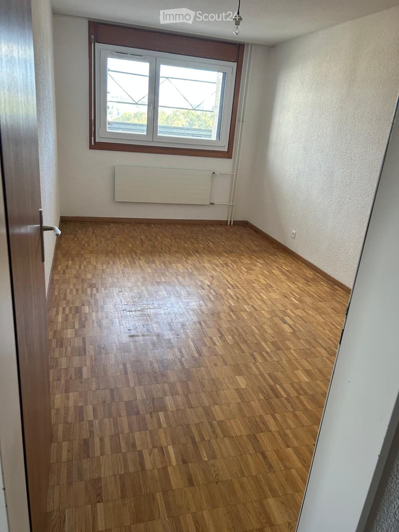 Empty room with wooden flooring, white walls, a window with a radiator below, and a visible door frame on the left side.