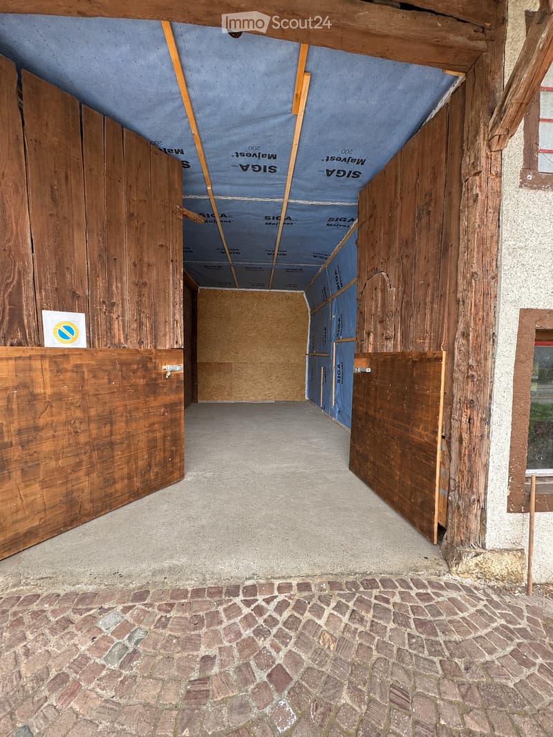 Empty corridor with exposed wooden beams, concrete floor, and cobblestone pathway. Wooden doors on both sides, with insulation visible in the ceiling area.