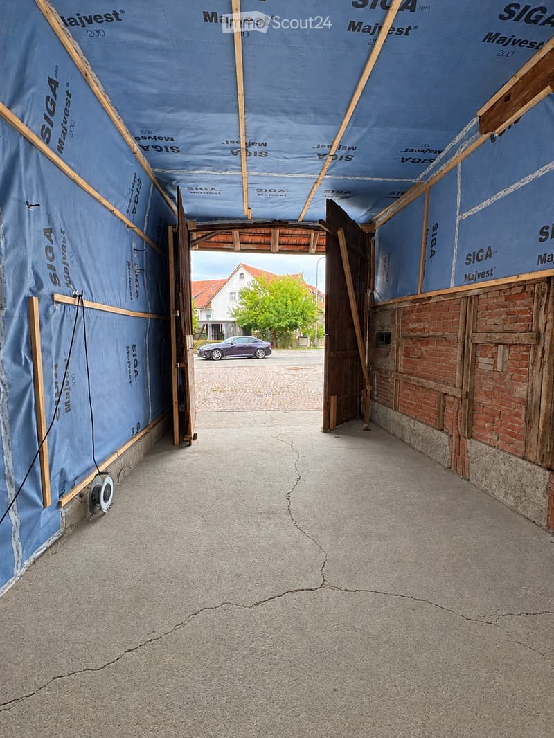 A covered corridor with blue protective tarps on the walls and ceiling, wooden framing, and a concrete floor. The corridor leads to an open area with a parked car and a building in the background.
