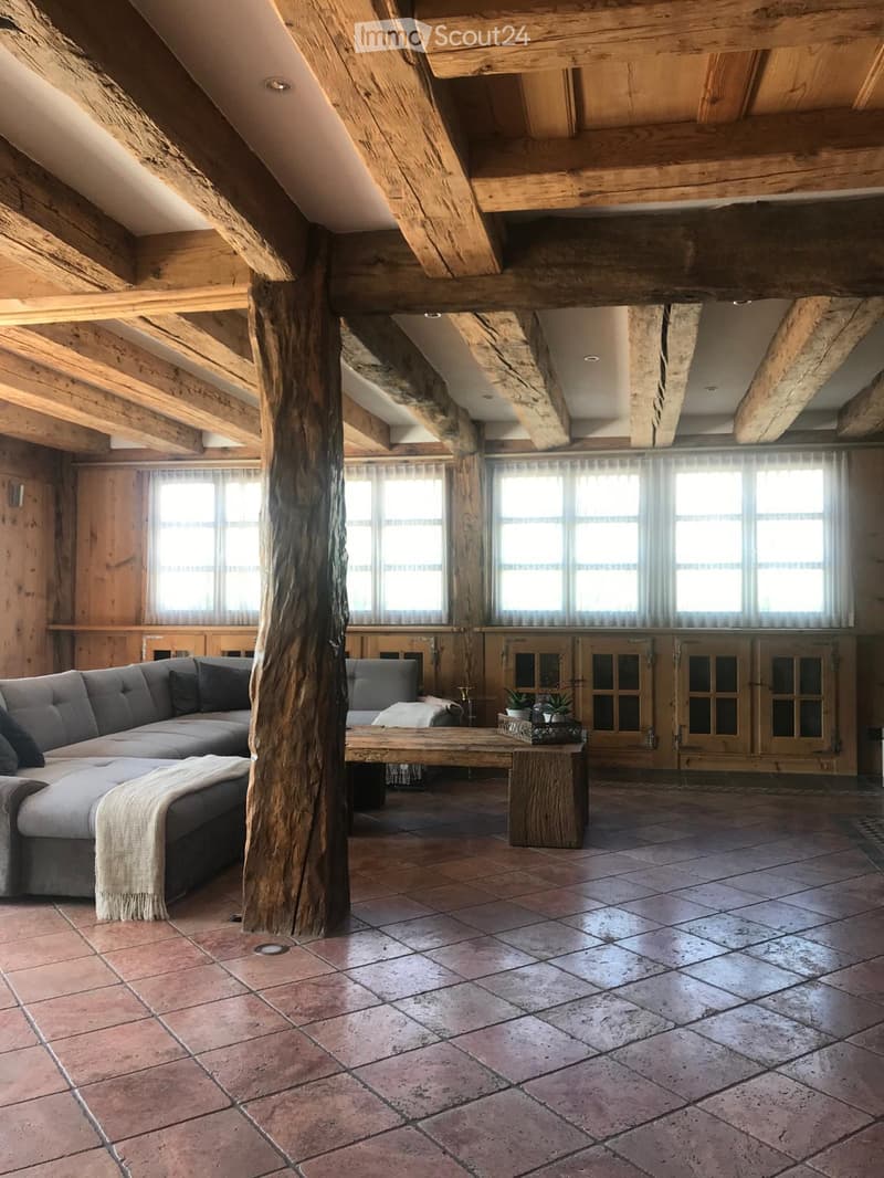 A room with wooden ceiling, sofa, center table, potted plants, and tiled floor.