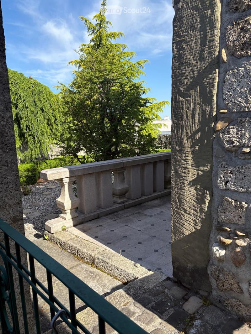 A balcony with stone pavement, stone railing, and green railing. The shadow of the railing reflects on the ground.