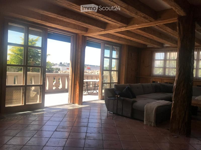 Large room with wooden beams, tiled floor, sectional sofa, table, large windows leading to balcony, table and chairs on balcony, beautiful view