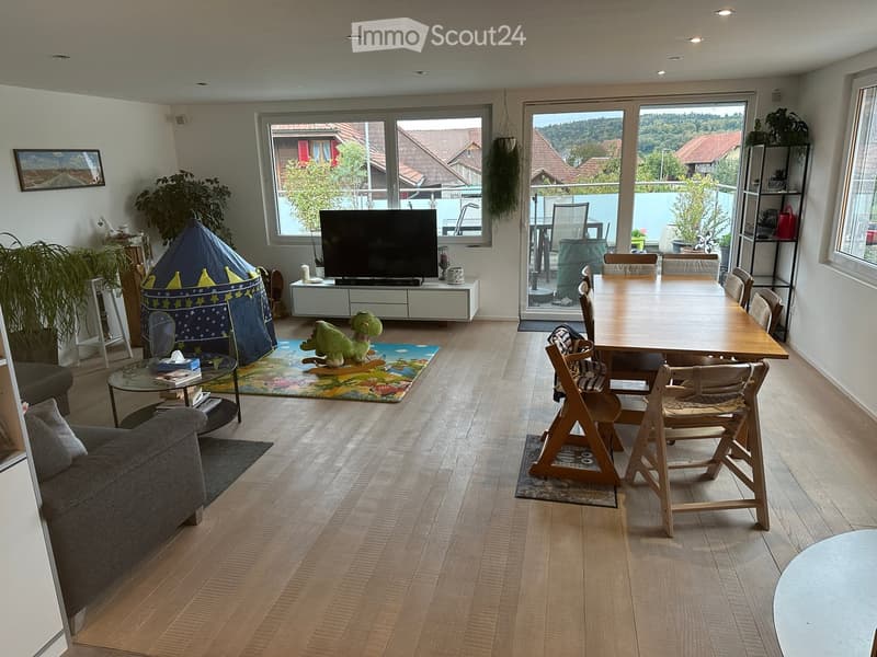 Open-plan living and dining area with wooden flooring, large windows, a sofa, coffee table, TV, dining table with chairs, and a play tent for children.