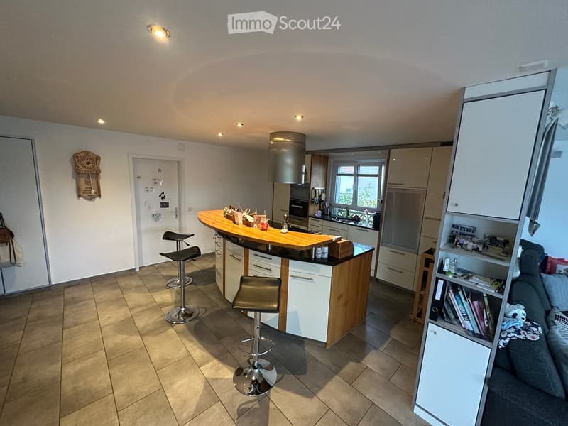Modern kitchen with a central island, bar stools, and built-in cabinets. The space features a tiled floor, recessed lighting, and a view through a window. A sofa and bookshelf are visible in the adjacent living area.