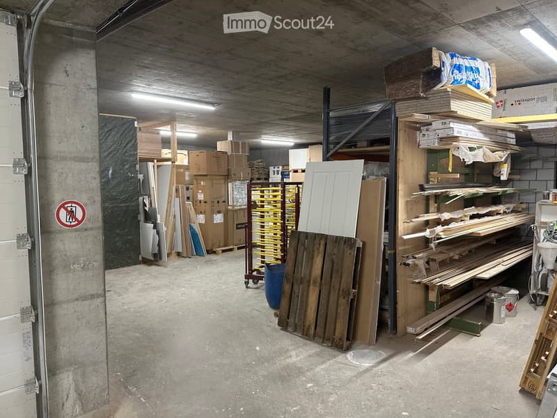 Empty storage area with concrete floors and walls, filled with shelves and stacks of wooden planks, cardboard boxes, and construction materials.