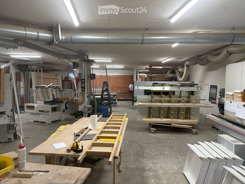 A spacious workshop with various woodworking tools and equipment, including a large worktable, saw, and a computer. The area is well-lit with fluorescent lighting and has exposed pipes and ductwork on the ceiling.