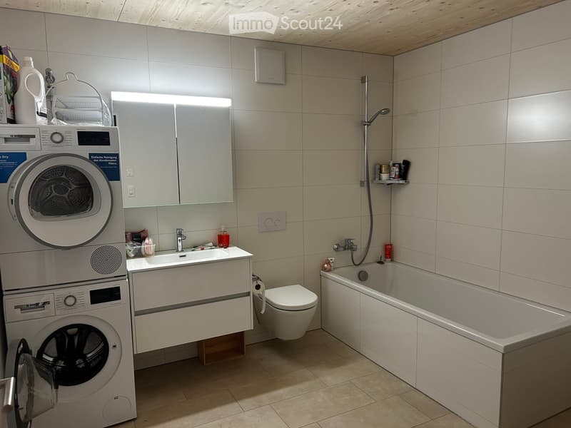 Modern bathroom with a washing machine, sink, toilet, bathtub, and mirror. The space features light-colored tiles and a sleek design.