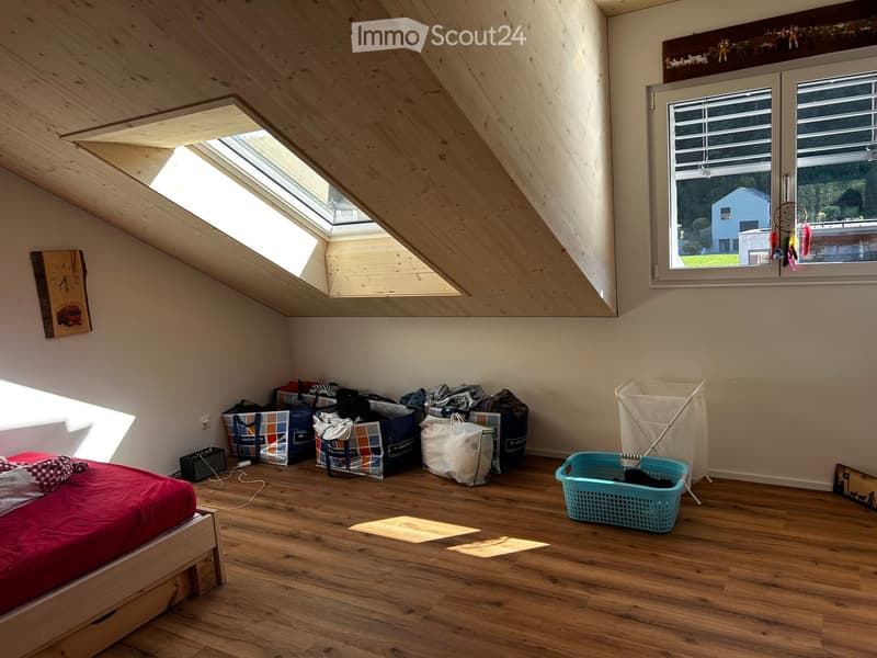 Bedroom with wooden floor, slanted wooden ceiling, skylight window, large window with blinds, bed with red blanket, laundry basket, laundry bag, and clock on wall.