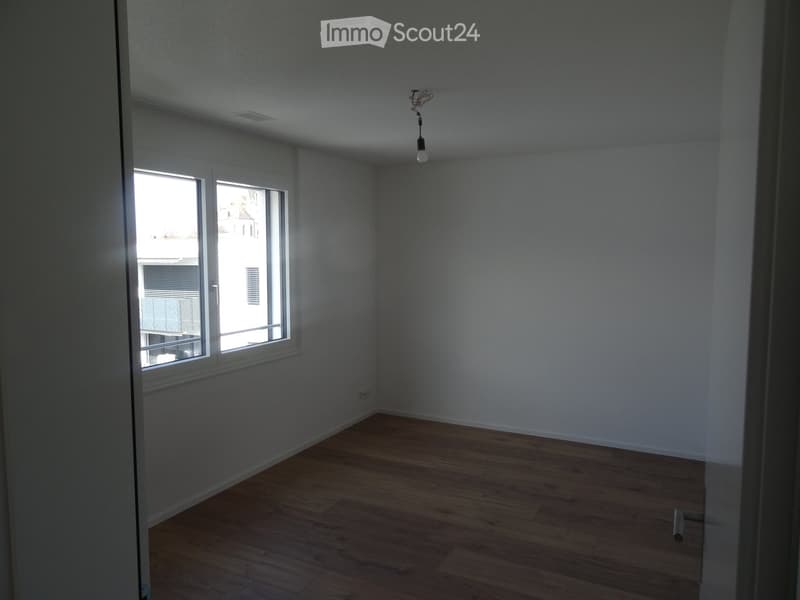 Empty room with wooden flooring, white walls, and a large window with blinds. The room has a single light fixture hanging from the ceiling.