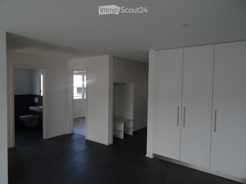 Empty room with white walls, dark tiled floor, built-in white wardrobe, and an open doorway leading to a small bathroom with a toilet and sink.