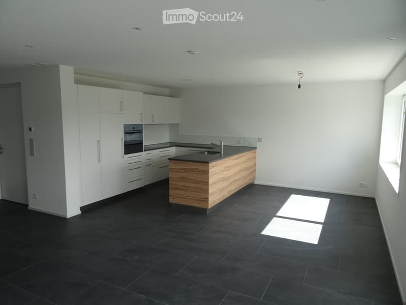 Modern kitchen with white cabinets, dark countertops, and a central island with wooden accents. The floor is dark tiled, and the space is well-lit with natural light from a window on the right.