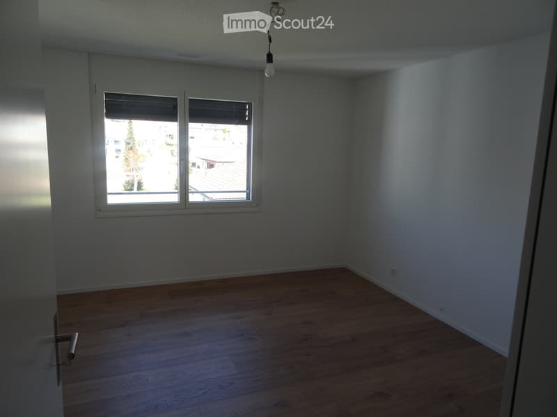 Empty room with light wooden flooring, white walls, and large windows offering a view of the outside area.