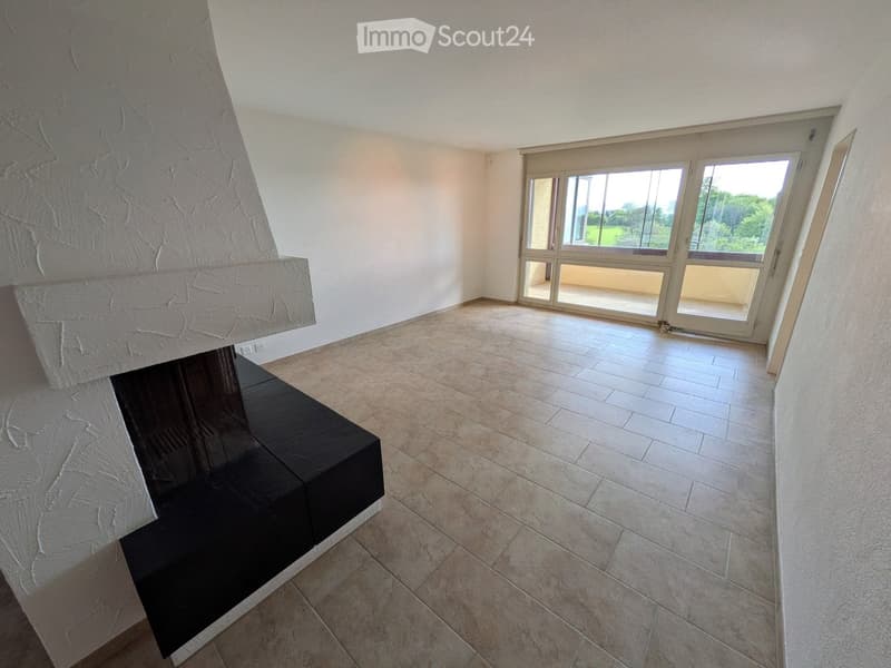 Empty room with a fireplace and large windows offering a view of greenery outside.