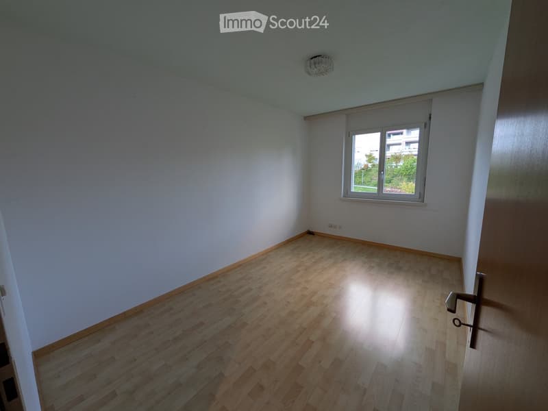 Empty room with light wooden flooring, white walls, and a large window letting in natural light.
