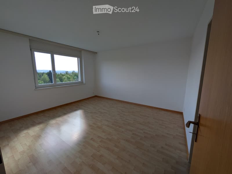 Empty room with light wooden flooring and large windows offering a scenic view of trees and landscape.