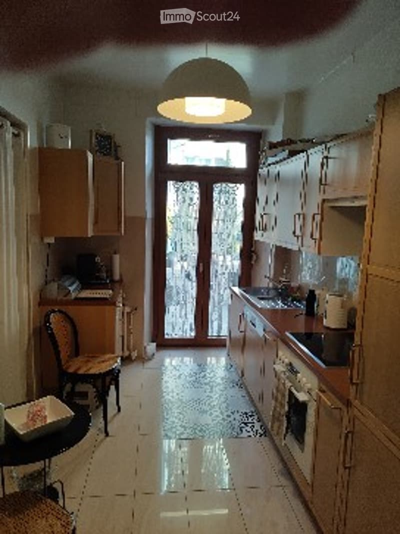 Bright kitchen with light wooden cabinets, a stove, sink, and a small dining area with a chair and table. A large glass door leads to an outdoor area.