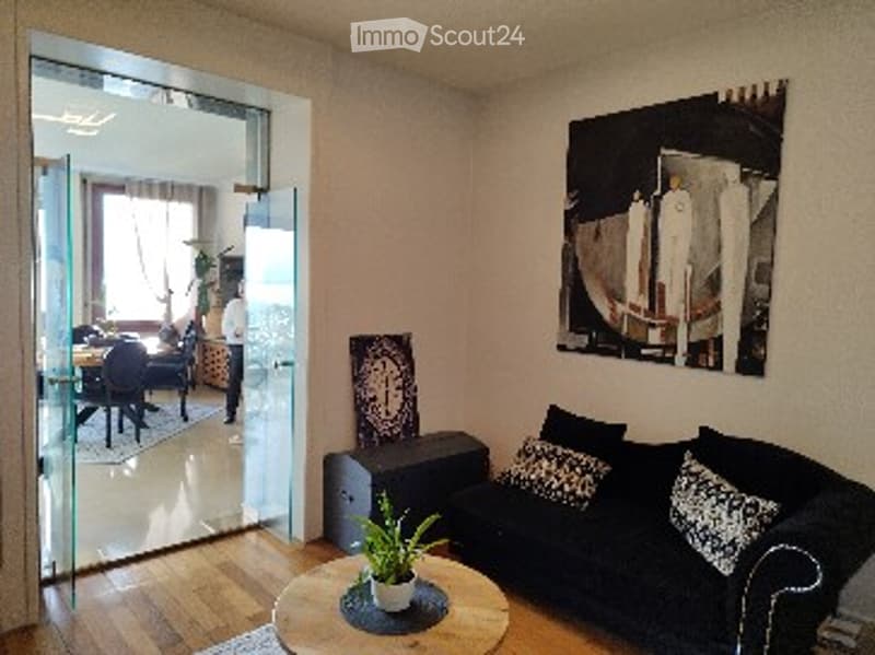 Modern living room with wooden flooring, large glass sliding door leading to another room, black sofa with patterned cushions, round coffee table with a potted plant, and a dining area visible through the doorway.