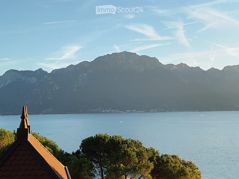 A scenic view of a lake with mountains in the background, featuring a small structure with a pointed roof in the foreground and trees scattered around.