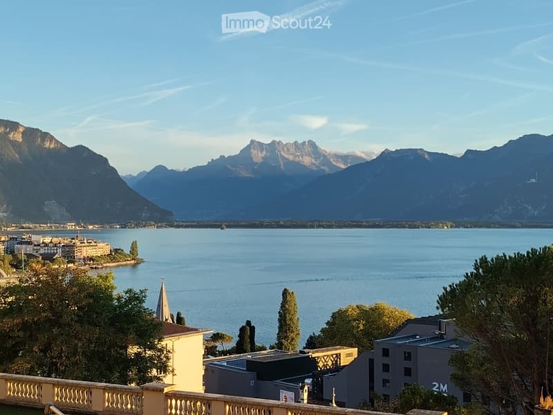 Panoramic view from a balcony overlooking a lake with distant mountains, featuring a town with buildings and trees in the foreground.