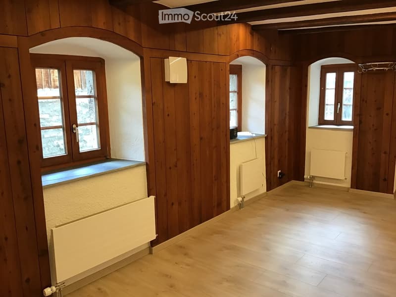 Empty room with wooden walls, wooden floor, and radiators. Windows with wooden frames and sills are visible.