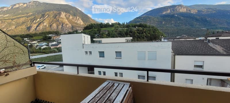 Balcony overlooking a town with white buildings and distant mountains, featuring a wooden bench and railing.